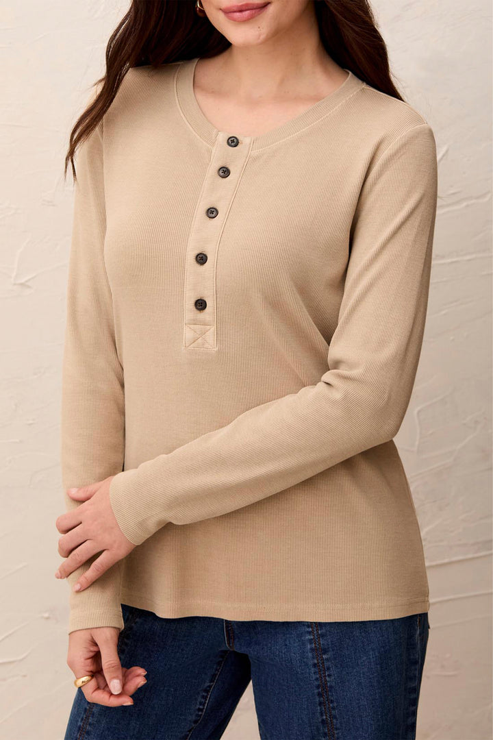 A woman wearing a beige long-sleeve henley top with dark buttons and blue jeans, standing against a light textured wall.