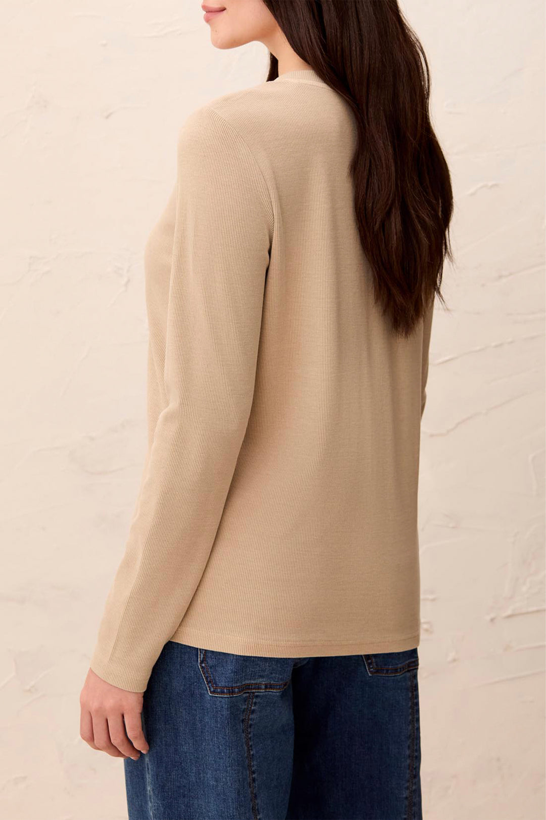 A woman shown from the back wearing a beige long-sleeve knit top and blue jeans against a light textured wall.