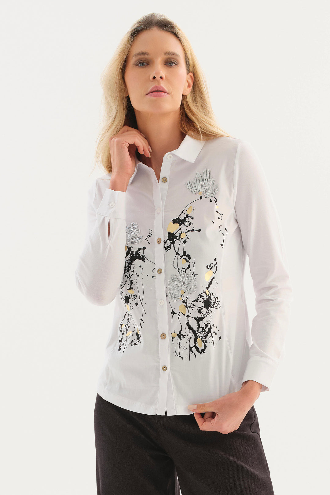 A woman modeling a white long-sleeve button-up shirt with a black, gray, and gold abstract brushstroke print on the front, paired with black pants, against a plain white studio background.