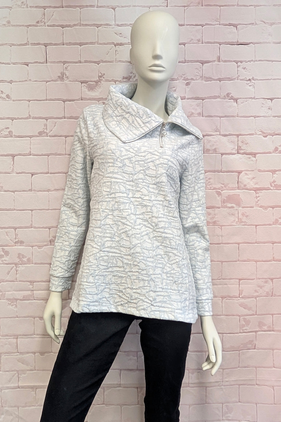 A mannequin wearing women's long-sleeve collared top with a textured, tonal abstract pattern in light gray and white, featuring a short silver zipper at the collar, shown on a light pink brick background