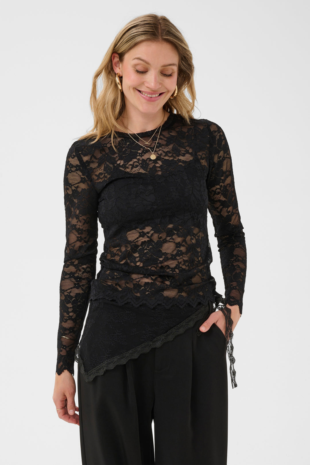 A woman wearing a black long-sleeve lace top with an asymmetrical scalloped hem and black pants, photographed against a light gray studio background.