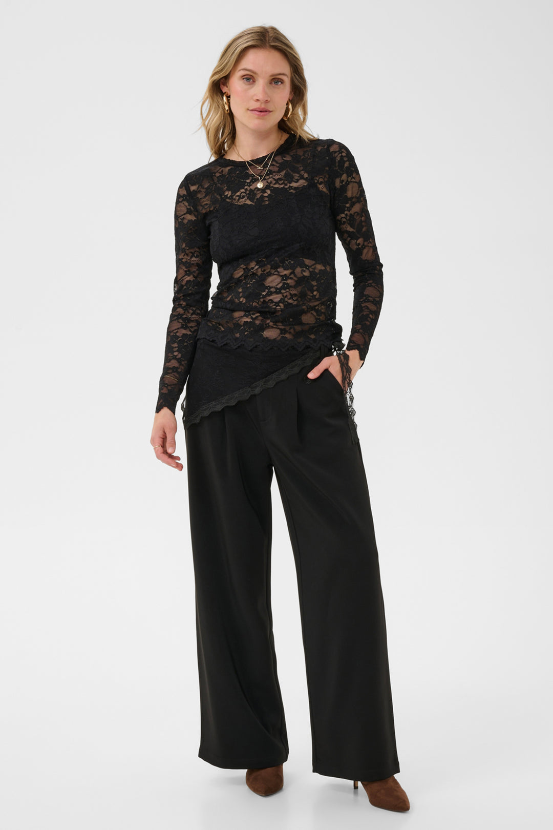 A woman modeling a black lace long-sleeve top with a scalloped hem, paired with wide-leg black trousers, against a plain light gray studio background.