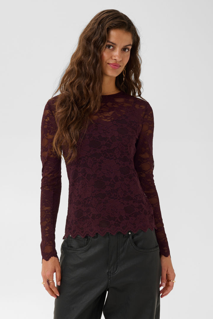 A woman wearing a burgundy brown sheer lace long-sleeve top and brown pants against a light gray studio background.