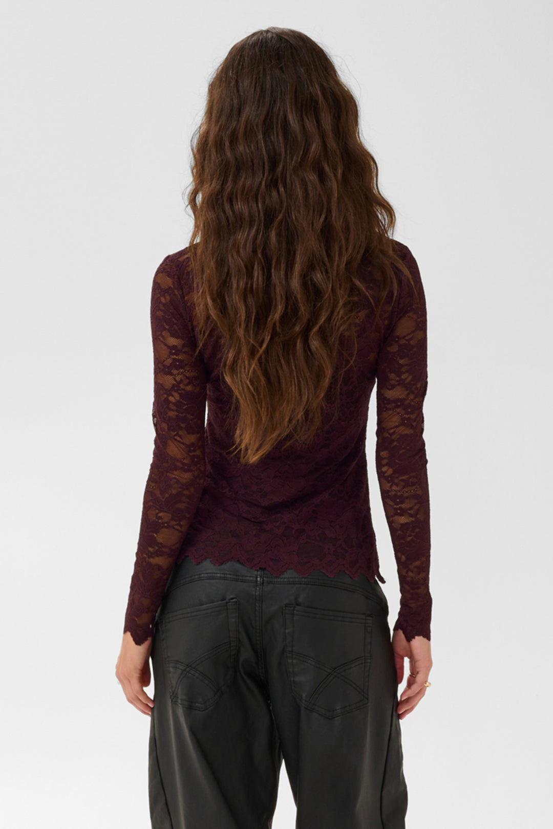 A woman shown from the back wearing a burgundy brown sheer lace long-sleeve top and brown pants against a light gray studio background.