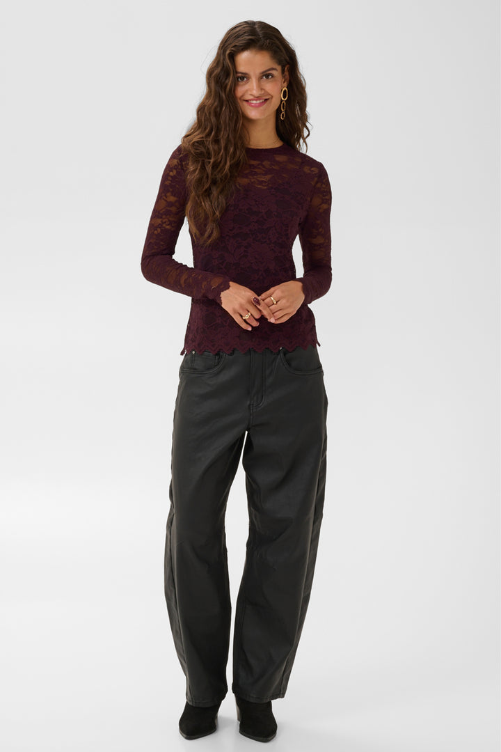 A woman wearing a burgundy brown sheer lace long-sleeve top and brown pants against a light gray studio background.