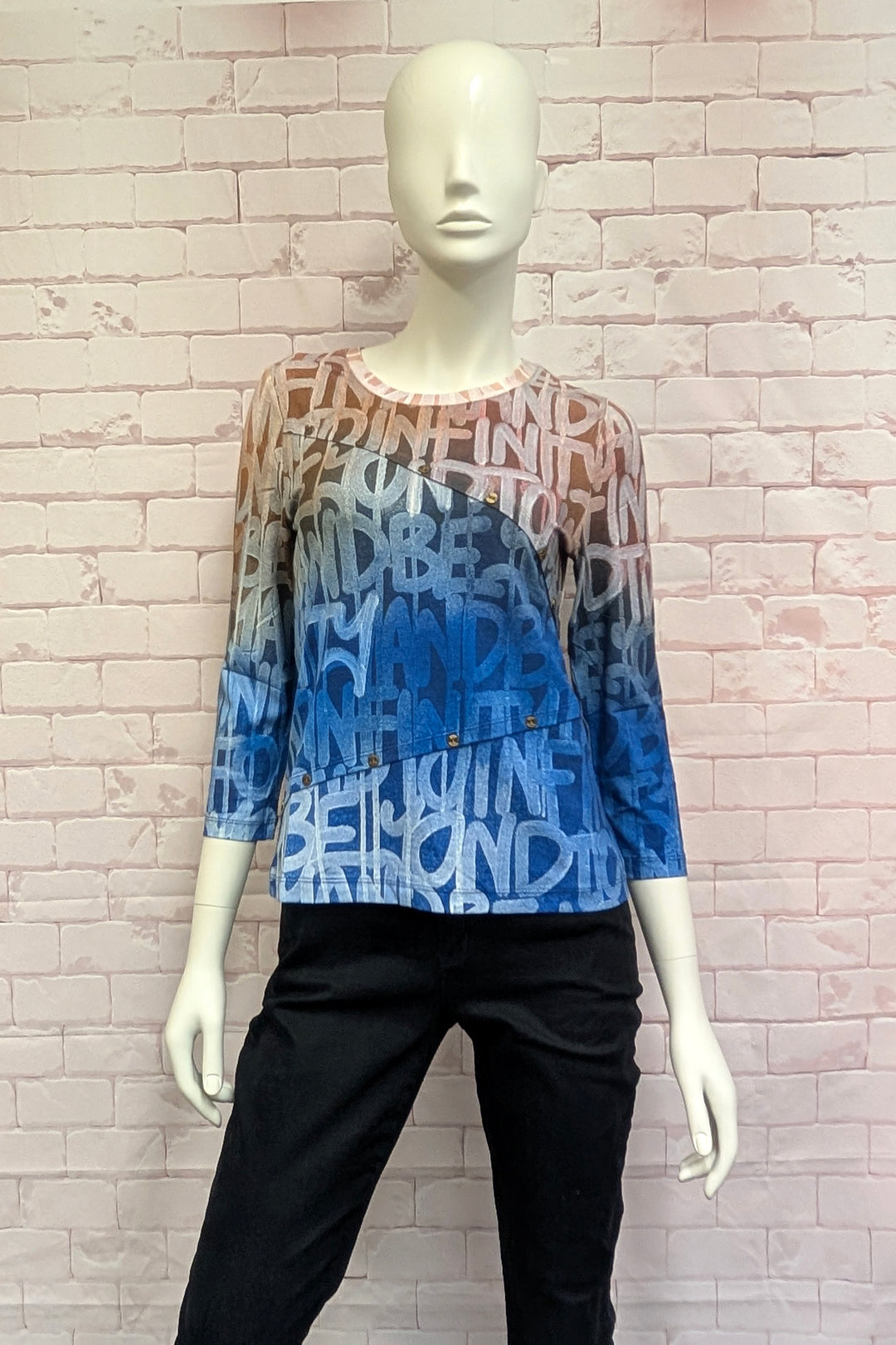 A women's mannequin wearing a three-quarter sleeve long-sleeve top with a gradient color transition from peach-orange at the shoulders to light blue and dark blue at the hem. The entire garment features an all-over abstract graffiti-style text pattern in white and blue.