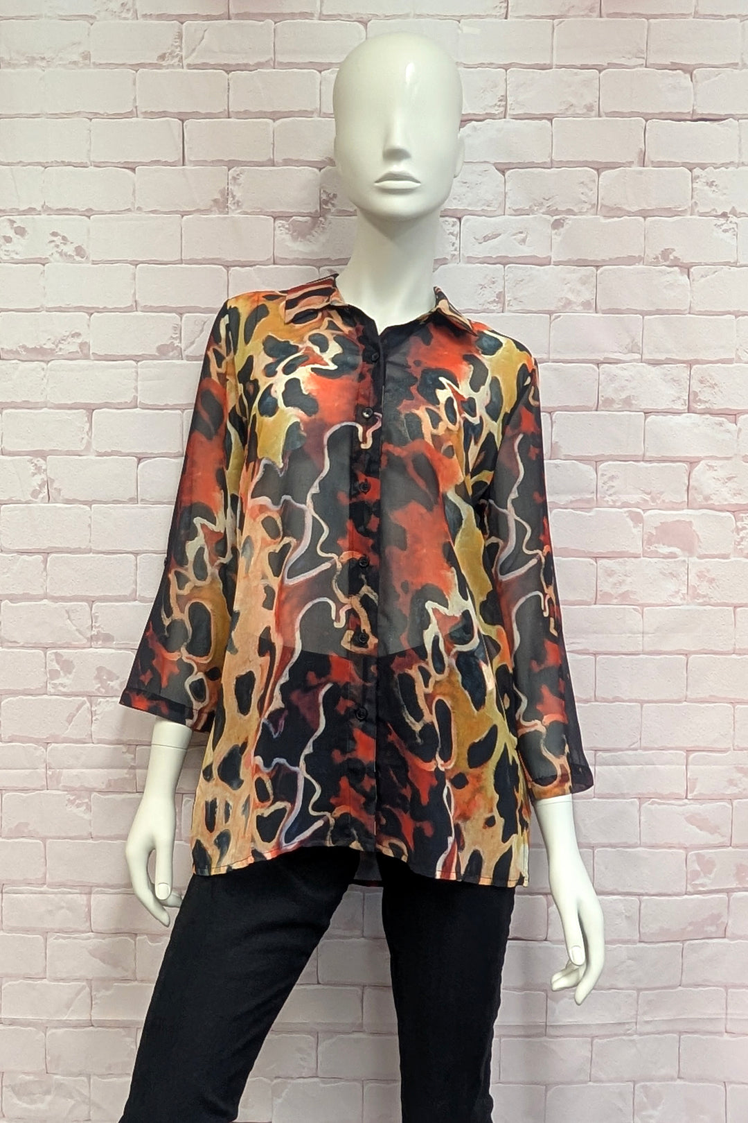 A three-quarter sleeve button-up blouse with an abstract wavy camouflage pattern in orange, black, and beige, displayed on a white mannequin against a light brick wall.