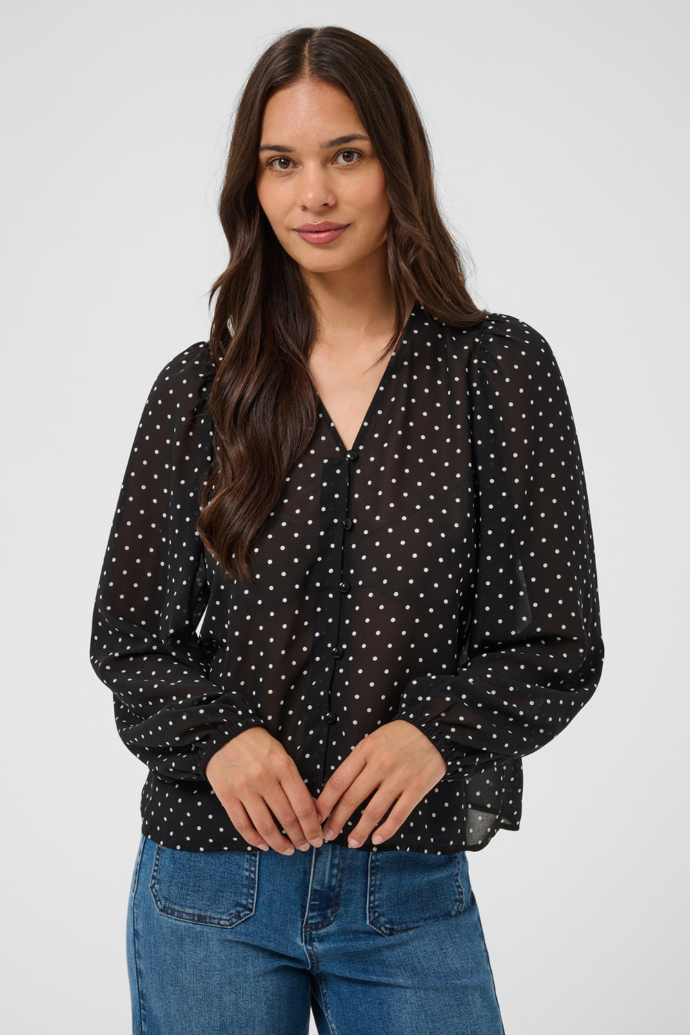 A woman standing in front of a light grey background wearing a black polka dot blouse and jeans. 
