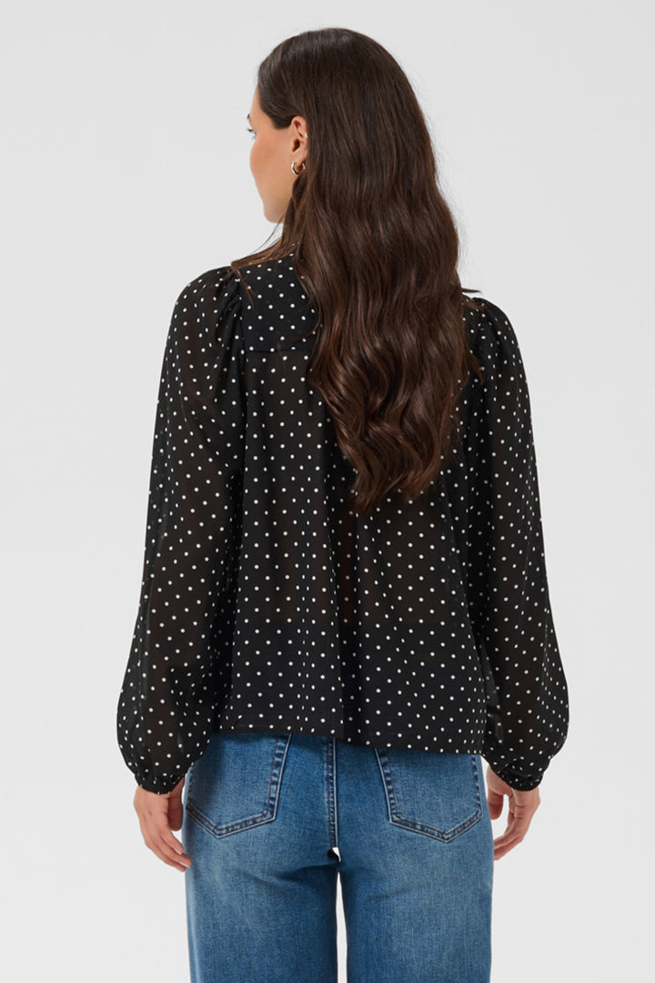 A woman shown from the back standing in front of a light grey background wearing a black polka dot blouse and jeans. 