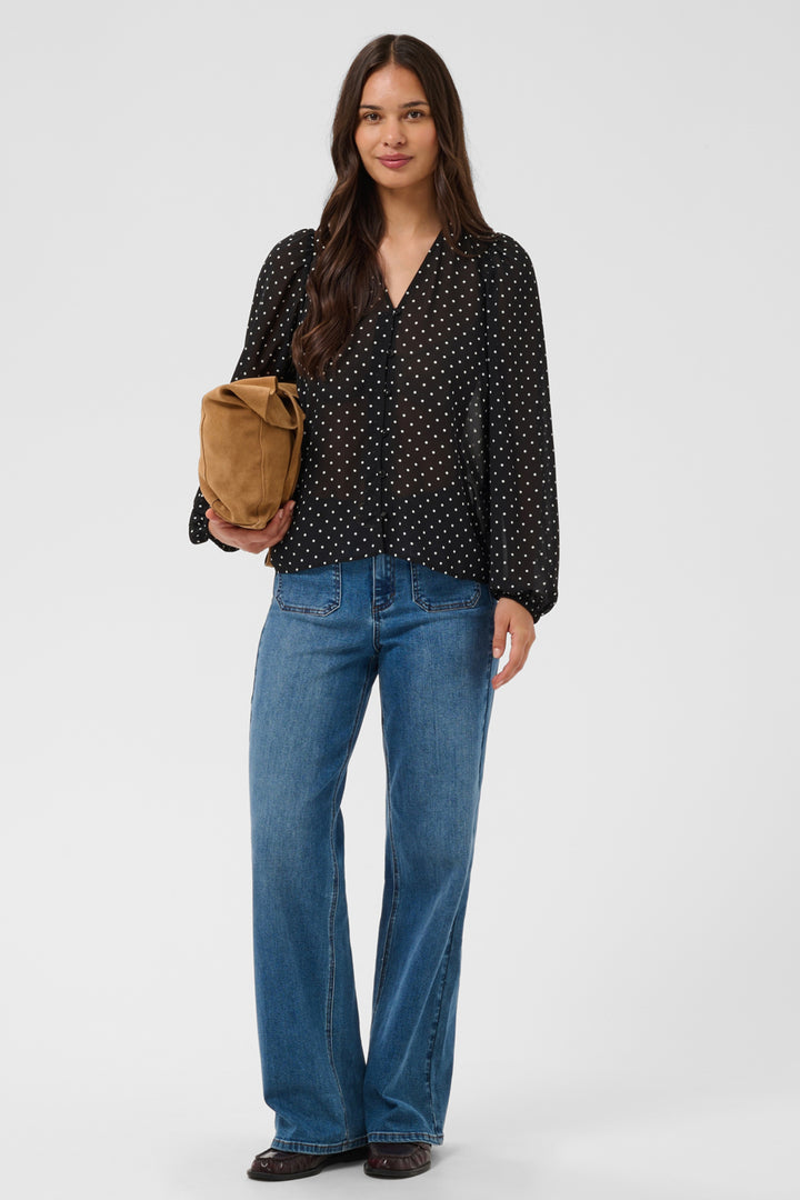 A woman standing in front of a light grey background wearing a black polka dot blouse and jeans. 