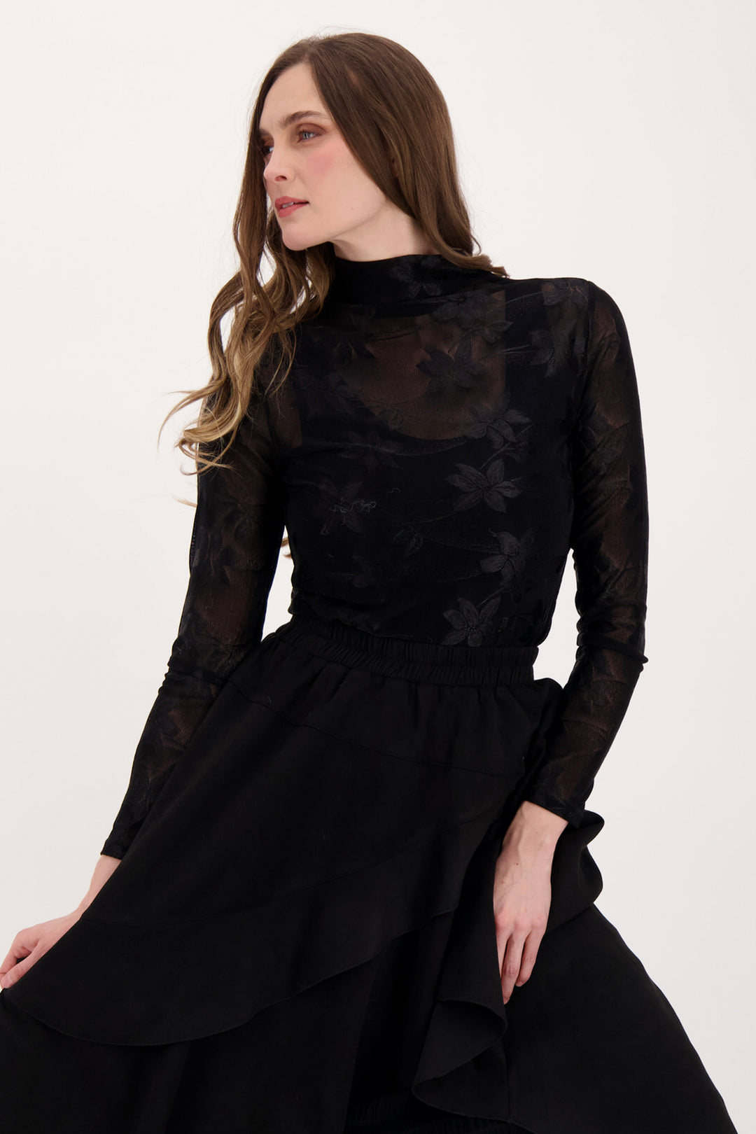 A woman modelling a sheer floral lace long sleeve top and black skirt in front of a white background. 