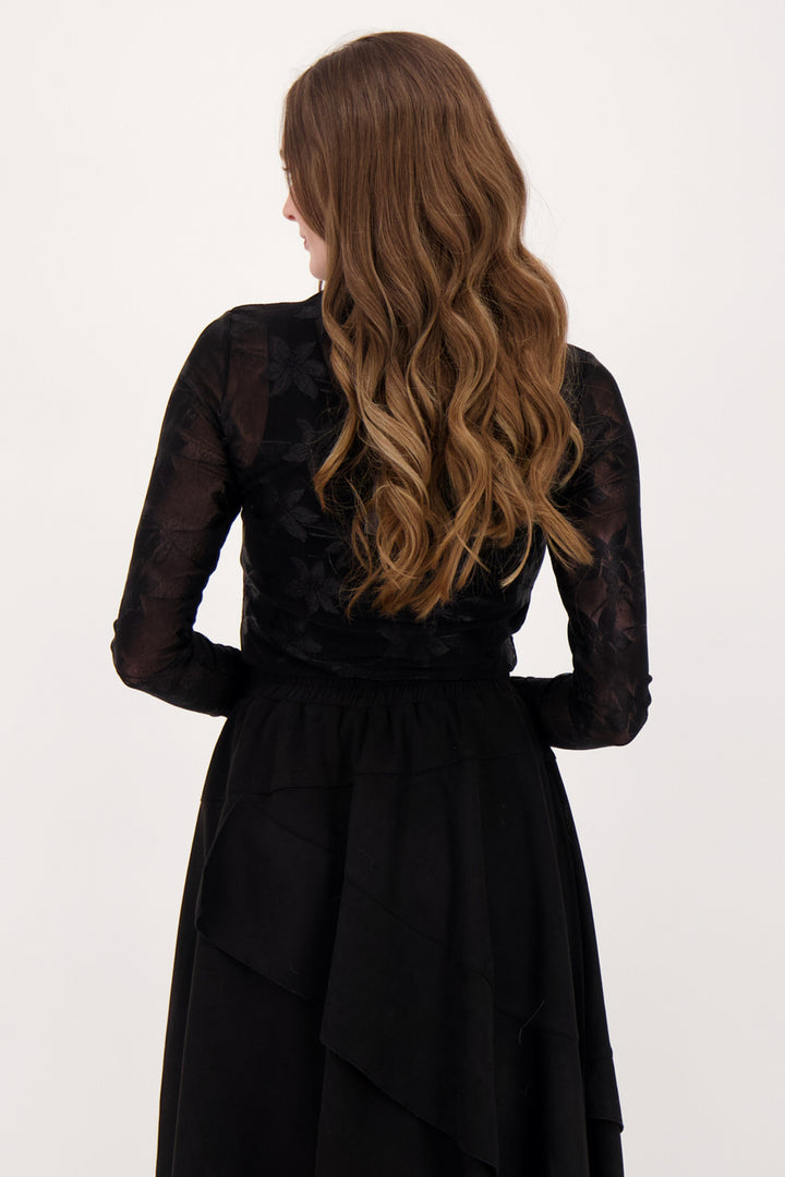 A woman modelling a sheer floral lace long sleeve top and black skirt in front of a white background. 