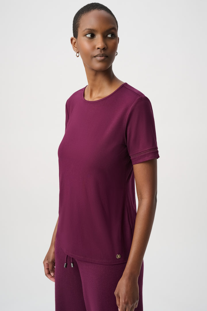 A woman modelling a burgundy short sleeve t-shirt standing in front of a light grey background. 