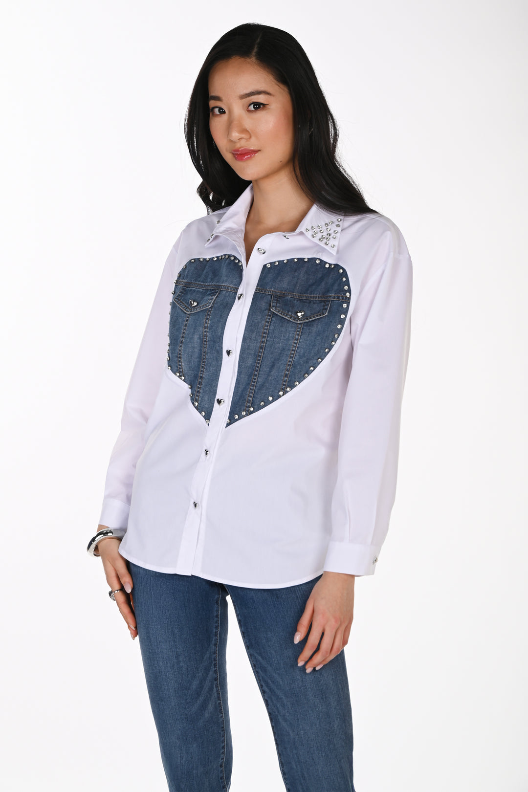 A woman wearing a white blouse with a denim heart graphic on the front and jeans standing in front of a white background. 
