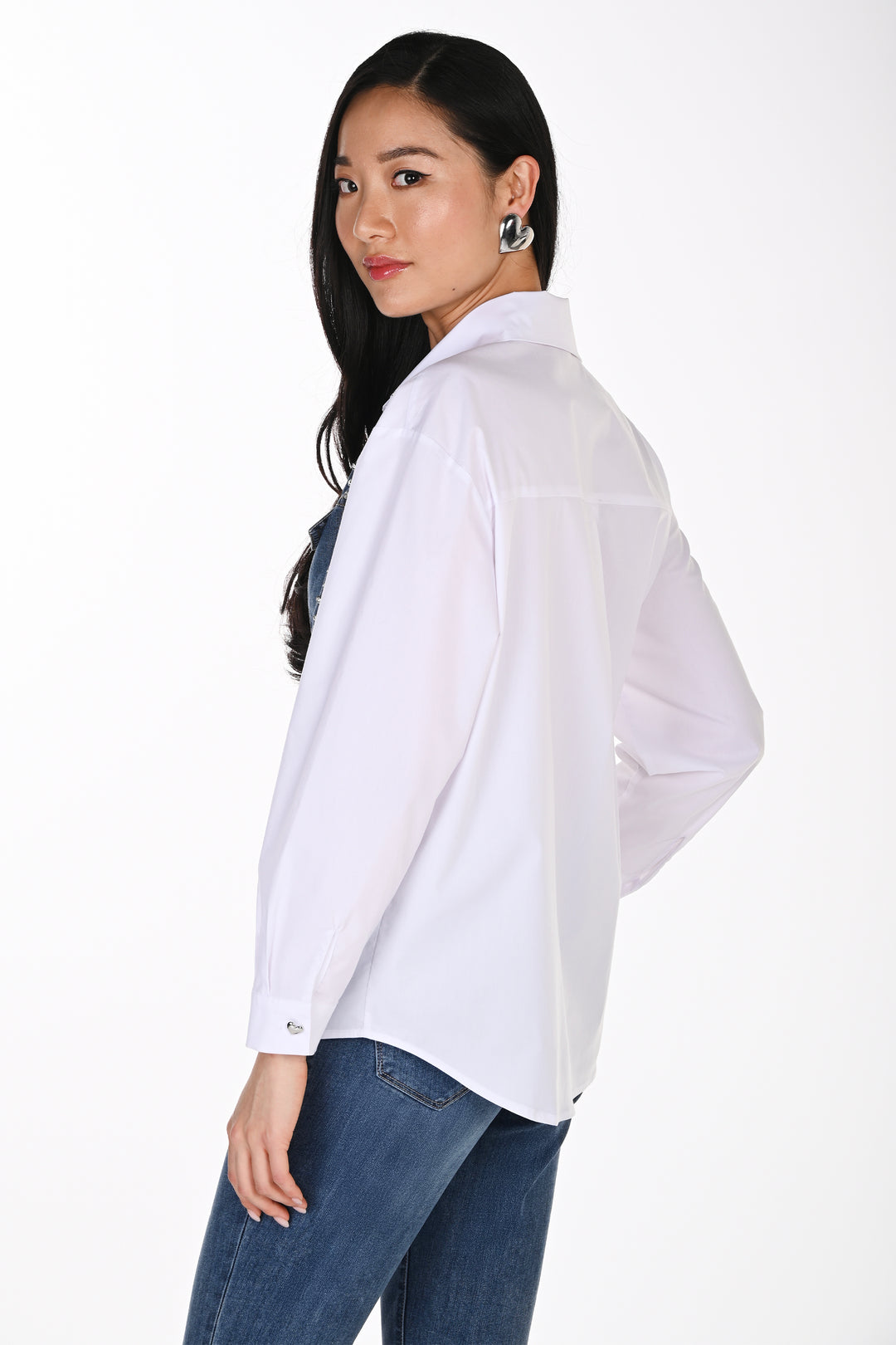 A woman shown from the back wearing a white blouse with a denim heart graphic on the front and jeans standing in front of a white background. 