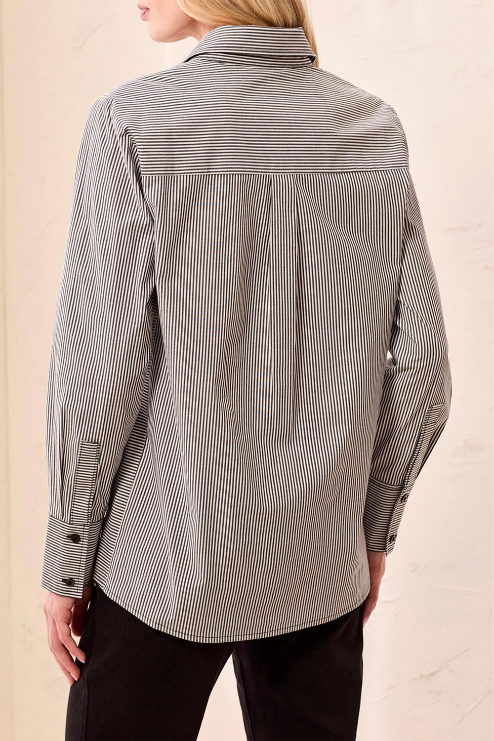 A woman shown from the back wearing a black and white long sleeve stripe blouse standing in front of a beige background. 