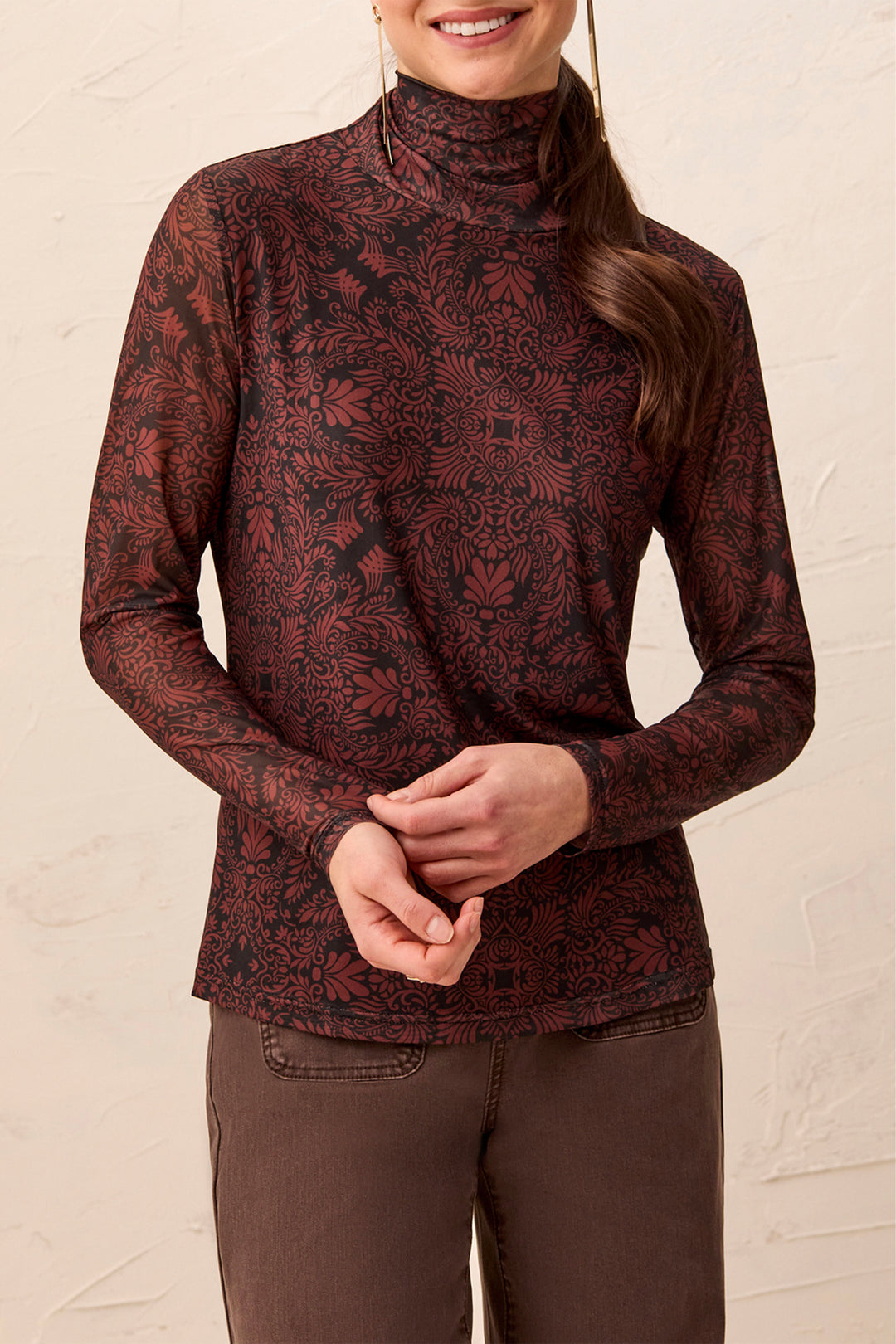A woman wearing a mock neck brown pattered long sleeve stop standing in front of a beige background. 
