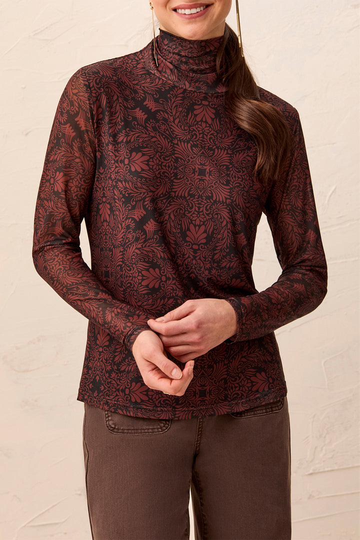 A woman wearing a mock neck brown pattered long sleeve stop standing in front of a beige background. 