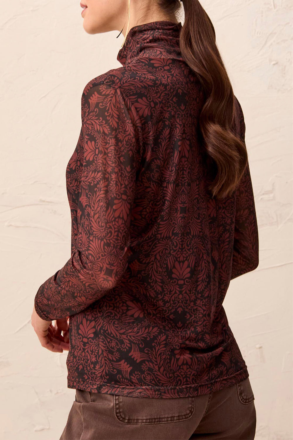 A woman shown from the back wearing a mock neck brown pattered long sleeve stop standing in front of a beige background. 