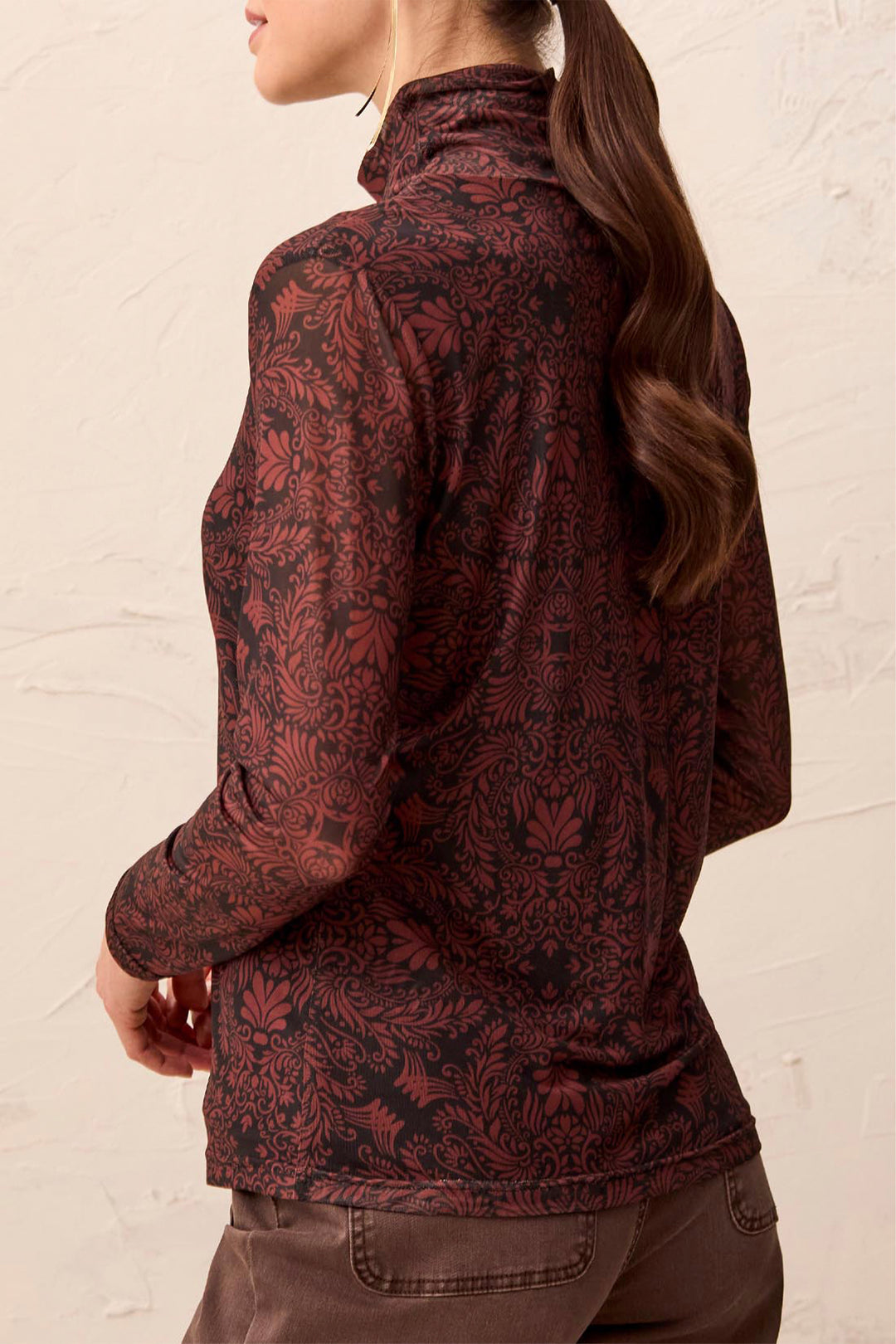 A woman shown from the back wearing a mock neck brown pattered long sleeve stop standing in front of a beige background. 