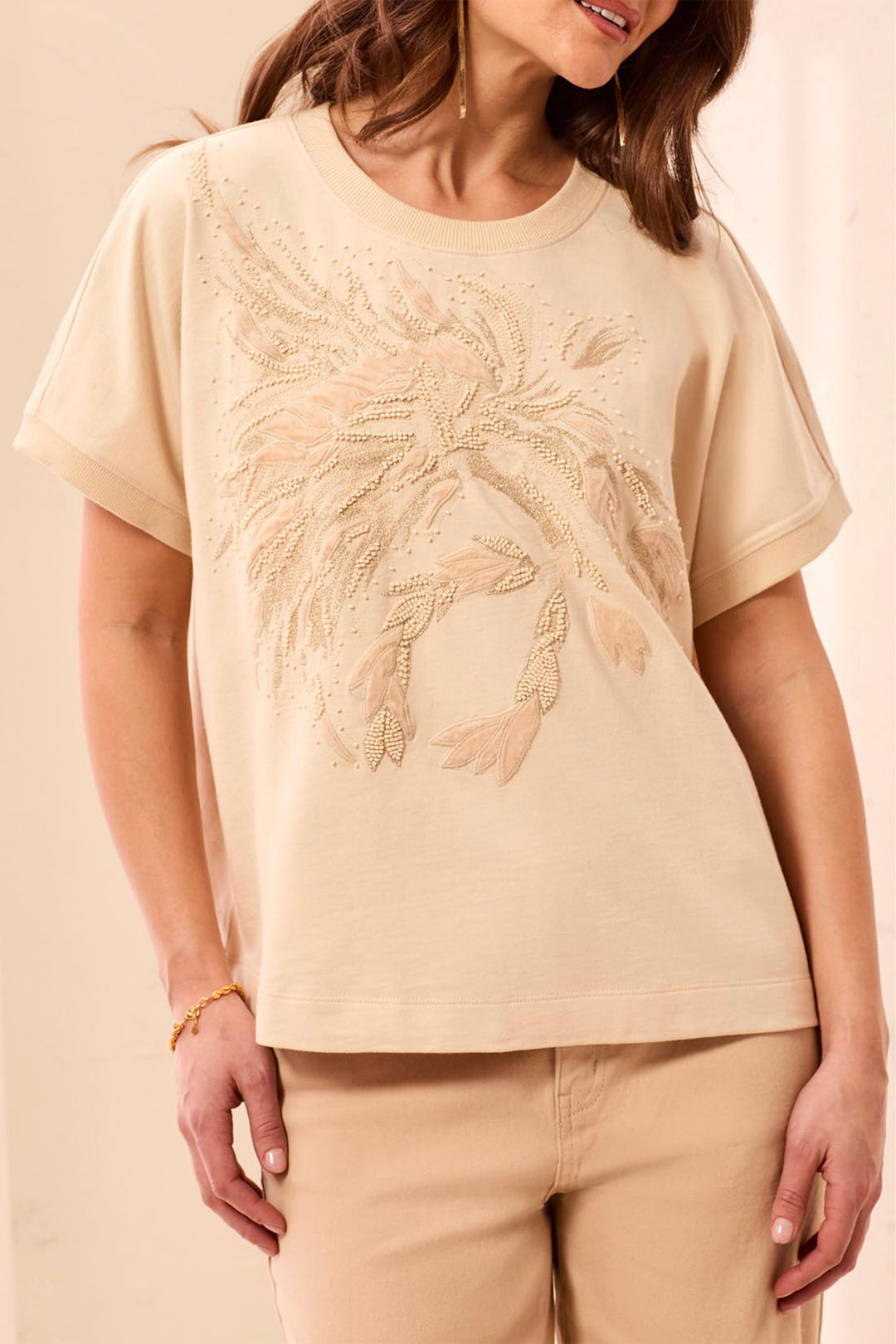 A woman wearing a cream bead and velvet embellished casual t-shirt standing in front of a beige background. 