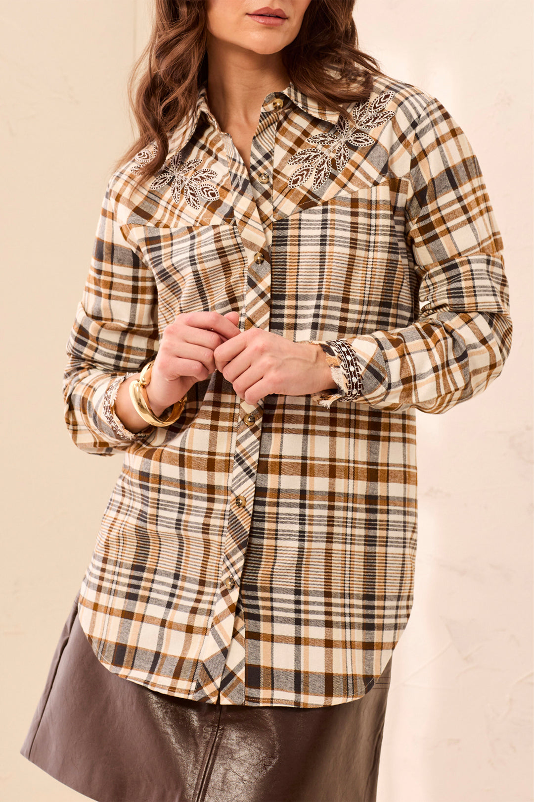 A woman wearing a beige and brown plaid long sleeve shirt standing in front of a beige background. 