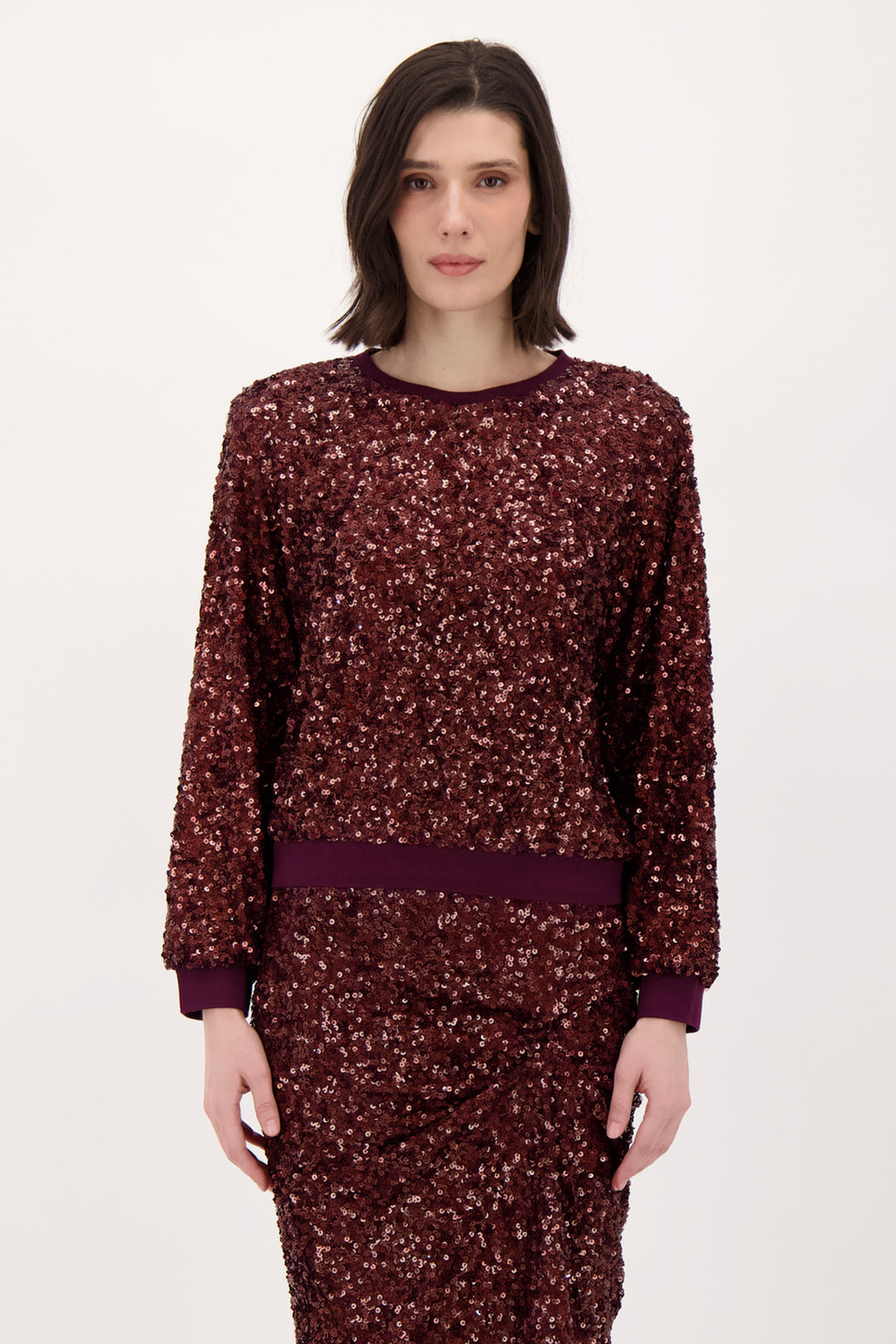 A woman wearing a long sleeve burgundy sparkly top and matching skirt standing in front of a light grey background. 