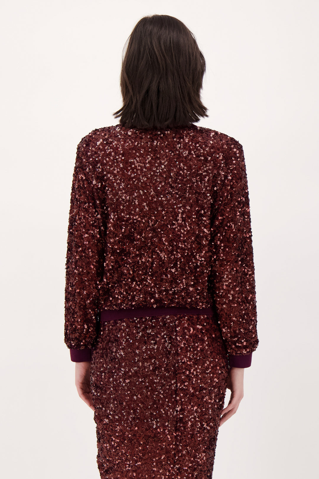 A woman shown from the back wearing a long sleeve burgundy sparkly top and matching skirt standing in front of a light grey background. 