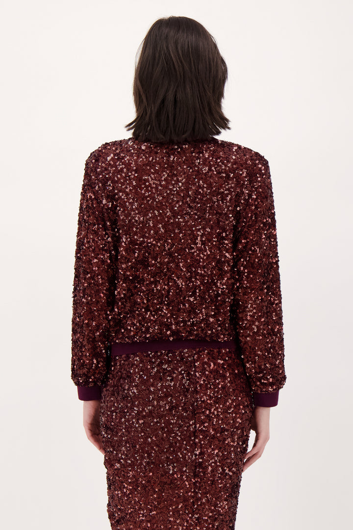 A woman shown from the back wearing a long sleeve burgundy sparkly top and matching skirt standing in front of a light grey background. 