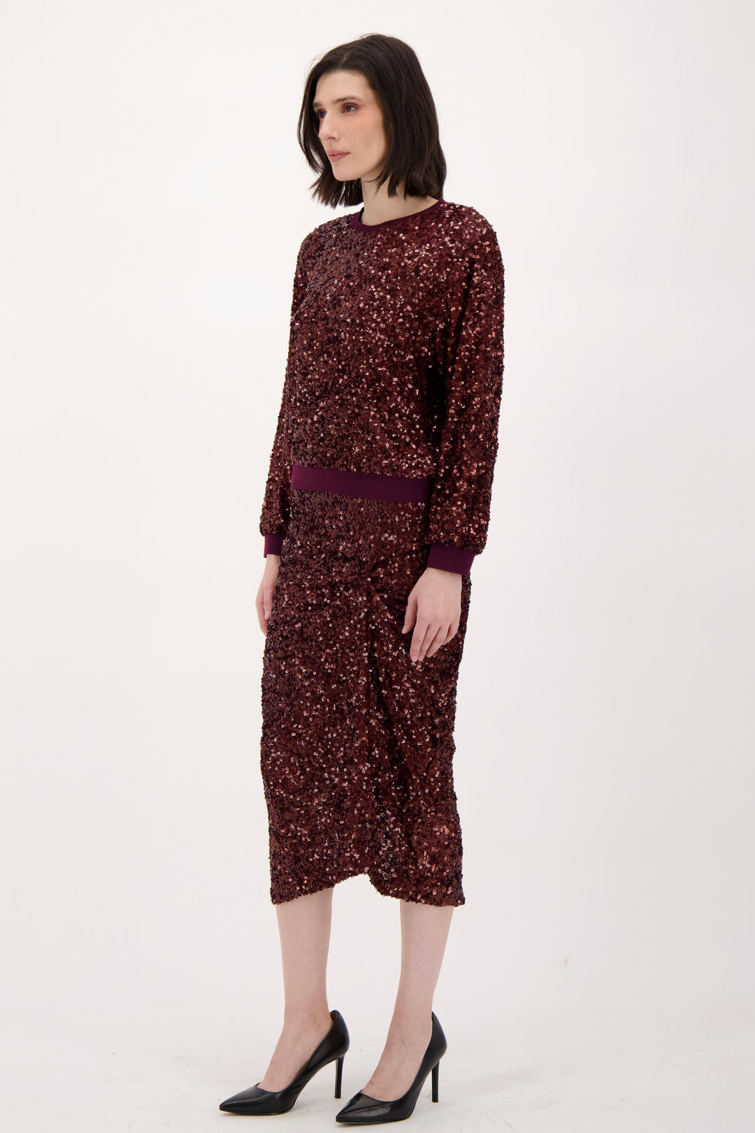 A woman wearing a long sleeve burgundy sparkly top and matching skirt standing in front of a light grey background. 