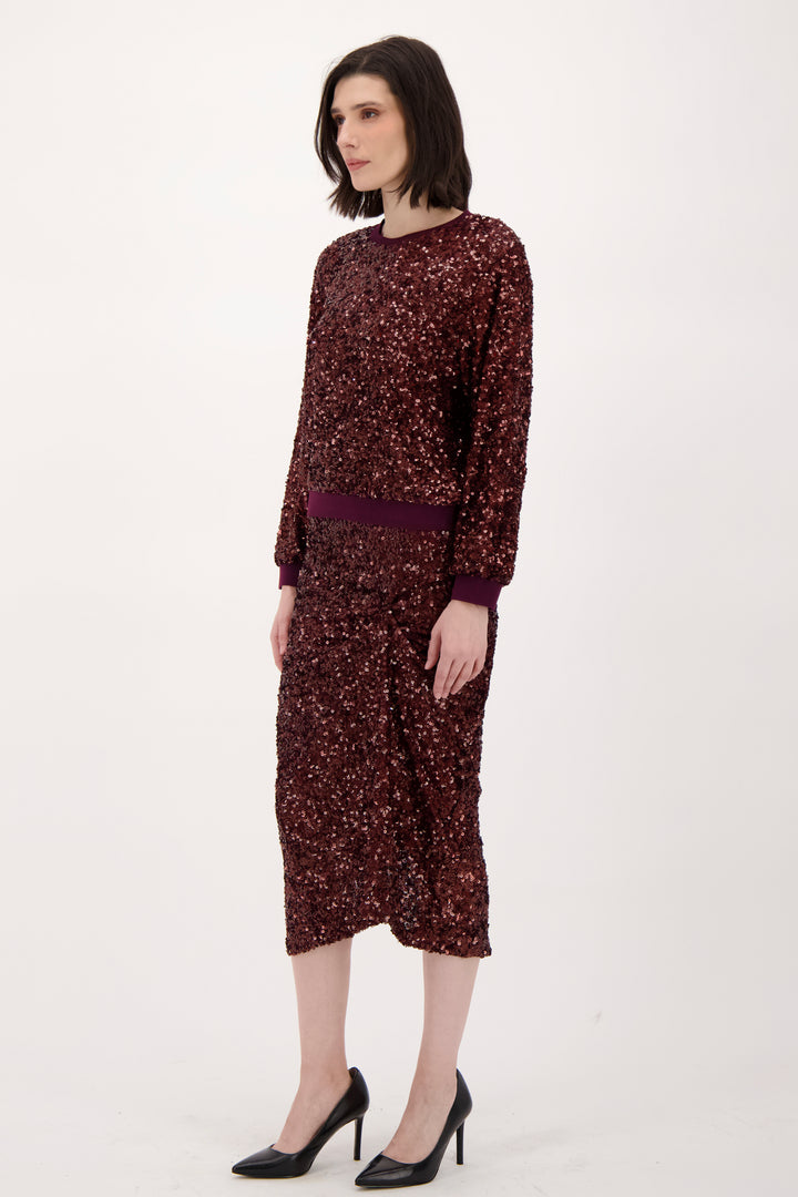A woman wearing a long sleeve burgundy sparkly top and matching skirt standing in front of a light grey background. 
