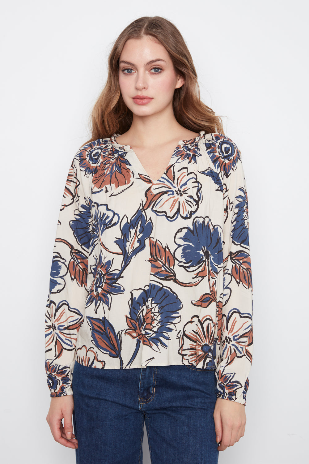A woman wearing a cream blouse with blue and brown flowers and long sleeves. She is wearing blue jeans and standing in front of a grey wall. 