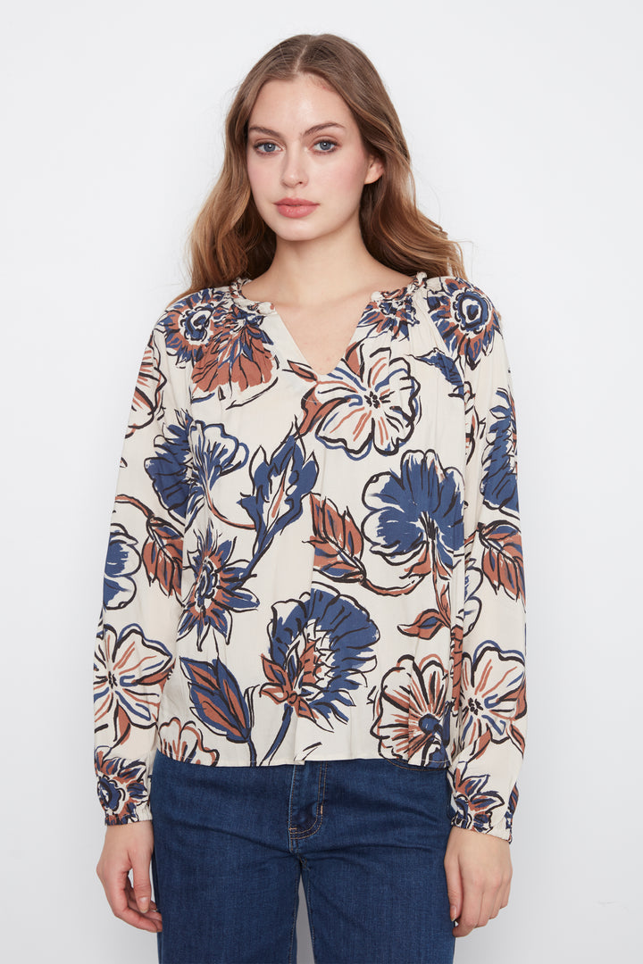 A woman wearing a cream blouse with blue and brown flowers and long sleeves. She is wearing blue jeans and standing in front of a grey wall. 