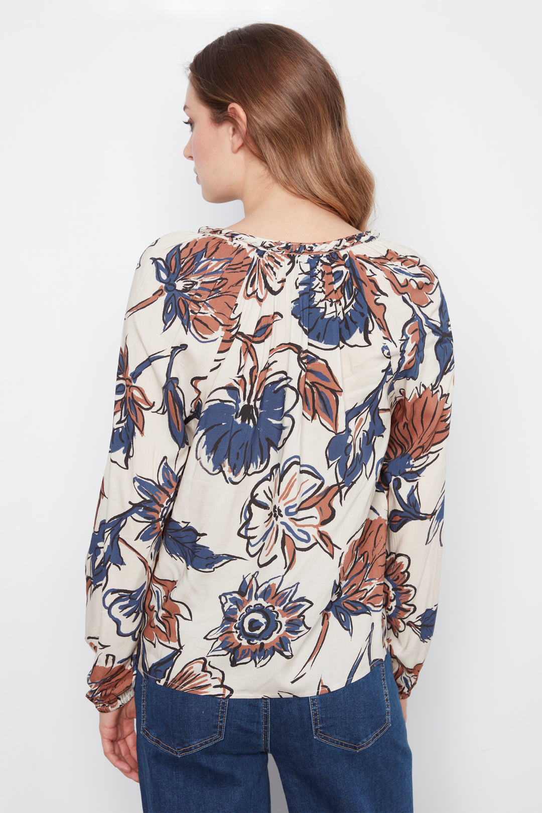 A woman shown from the back wearing a cream blouse with blue and brown flowers and long sleeves. She is wearing blue jeans and standing in front of a grey wall. 