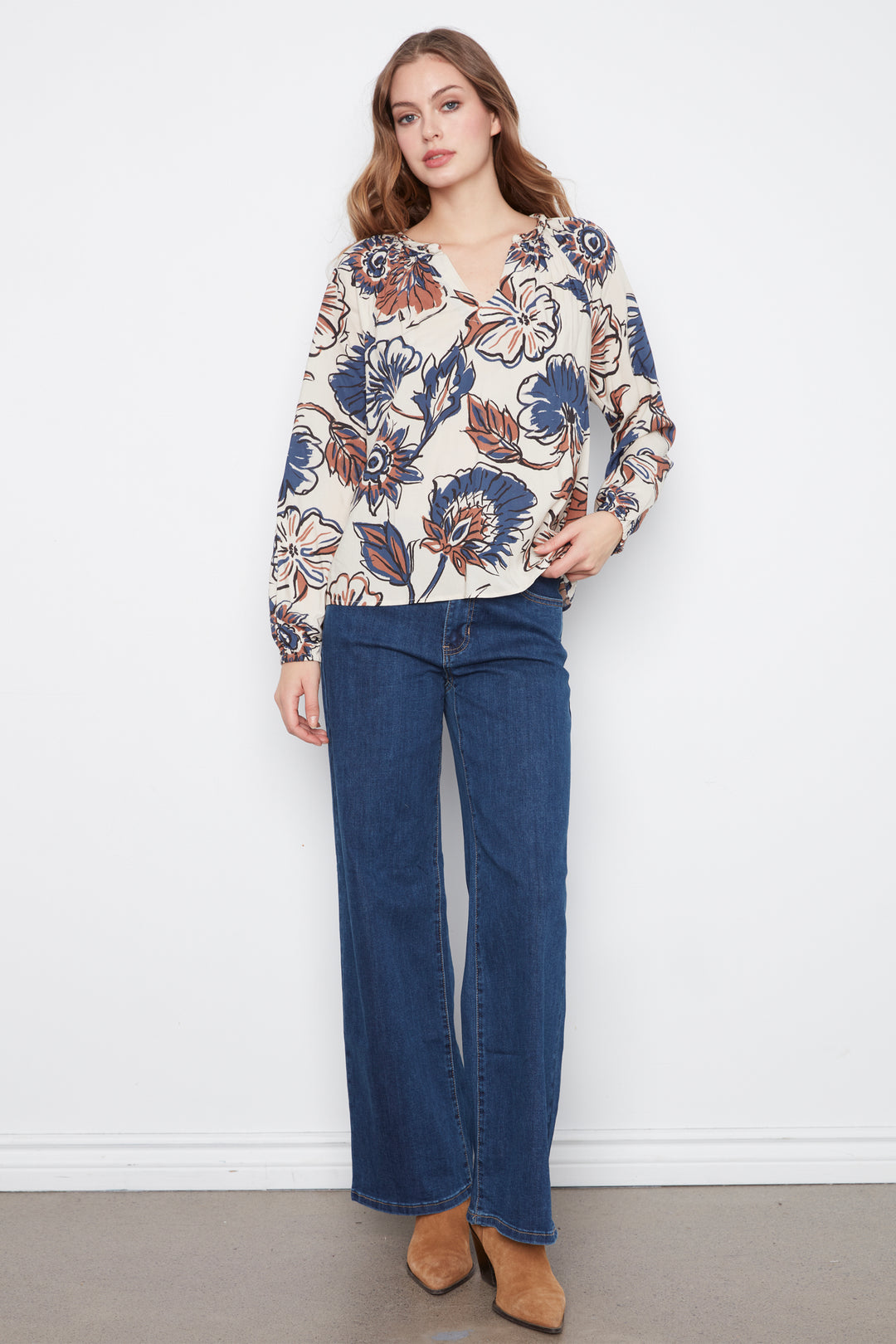 A woman wearing a cream blouse with blue and brown flowers and long sleeves. She is wearing blue jeans and standing in front of a grey wall. 