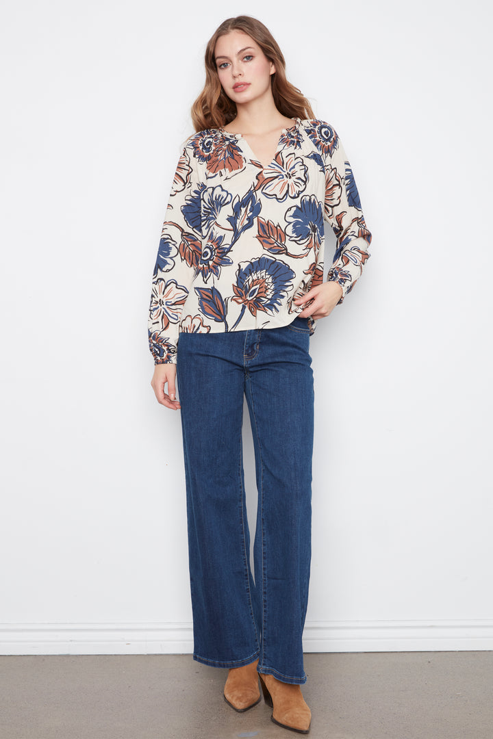 A woman wearing a cream blouse with blue and brown flowers and long sleeves. She is wearing blue jeans and standing in front of a grey wall. 
