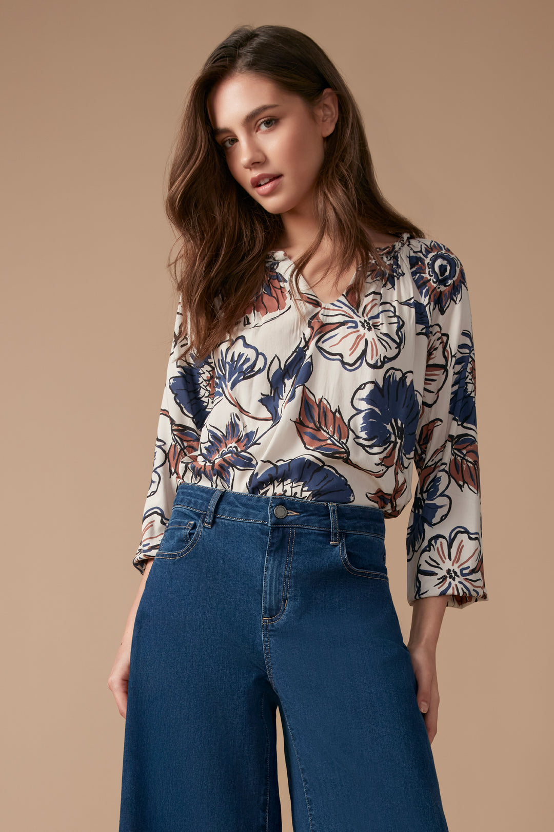 A woman wearing a cream blouse with blue and brown flowers and long sleeves. She is wearing blue jeans and standing in front of a beige wall. 