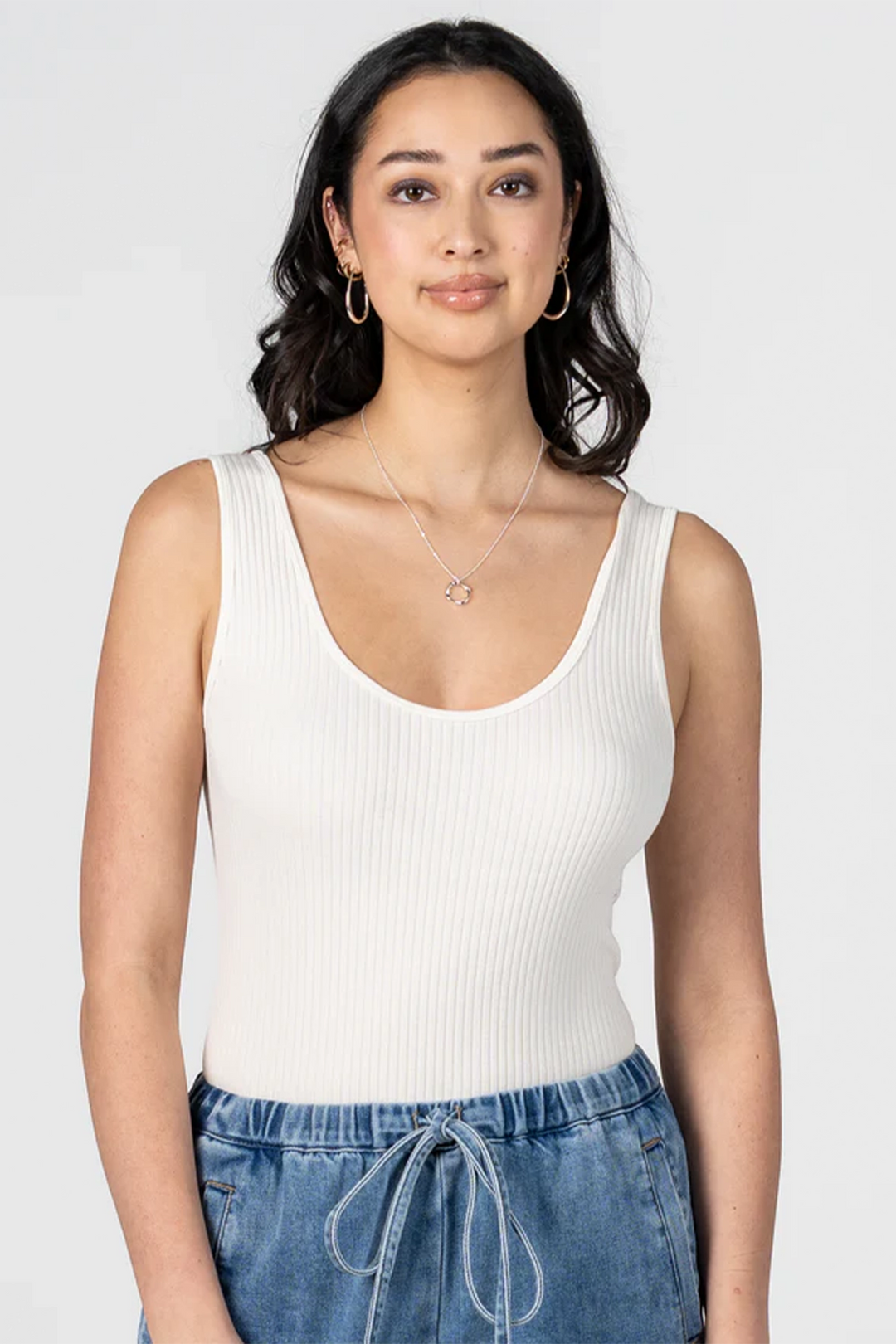 Woman wearing an ivory white tank top and blue jeans against a plain background