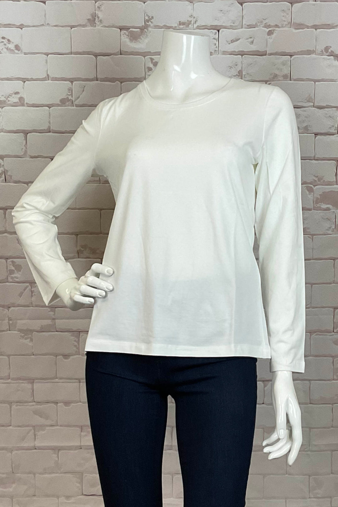 A long sleeve white top displayed on a mannequin with a crew neckline, made from a soft fabric, presented against a brick wall background.