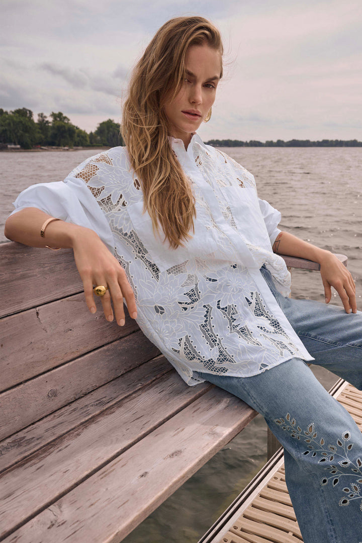 Woman sitting on a wooden bench by a body of water wearing a white lace embroidered blouse and blue jeans.