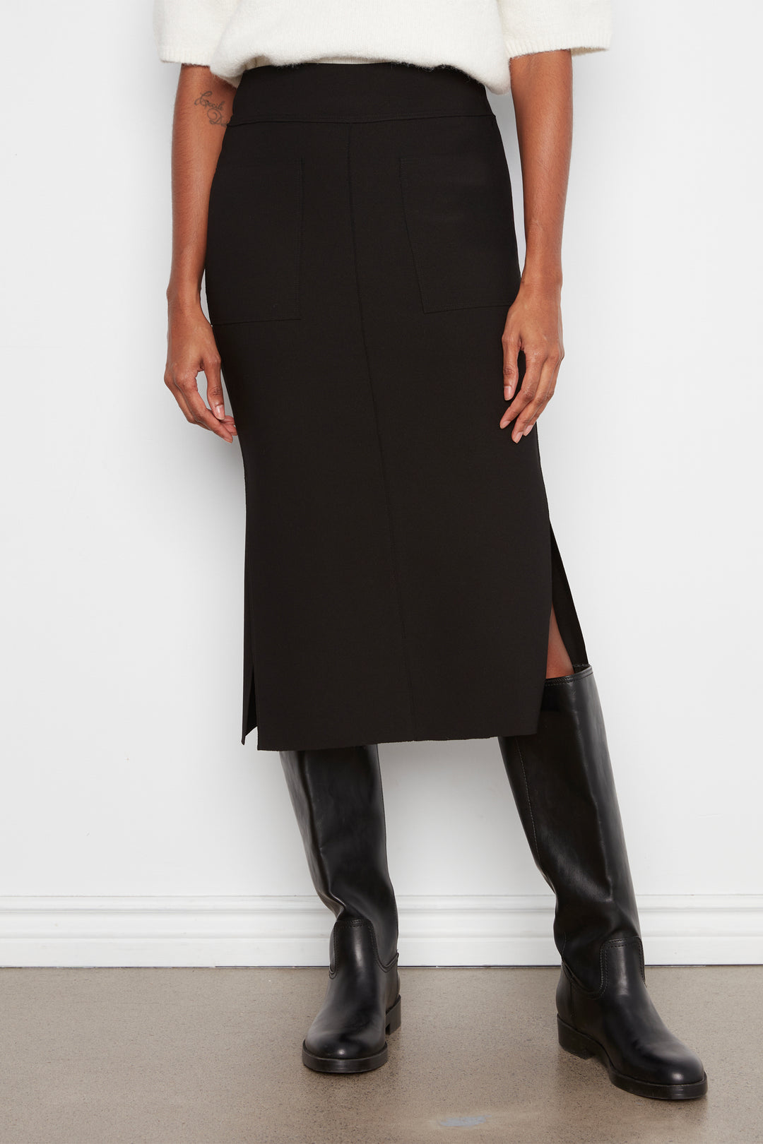 A model wearing a black midi skirt with a side slit, styled with a white top and black knee-high boots against a white wall.