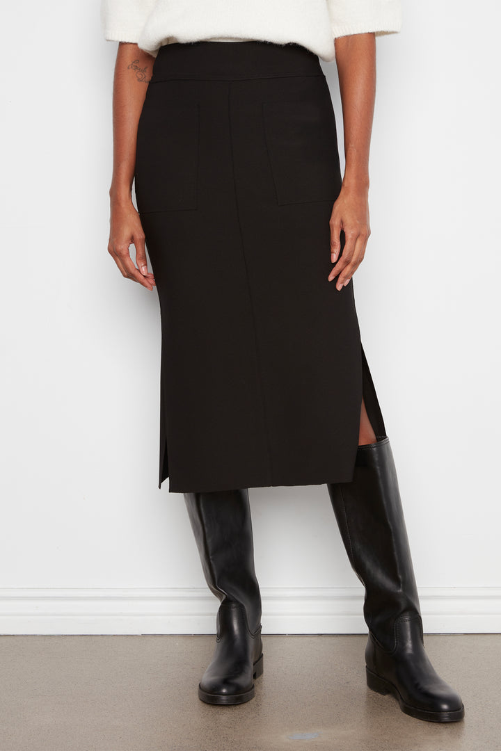 A model wearing a black midi skirt with a side slit, styled with a white top and black knee-high boots against a white wall.