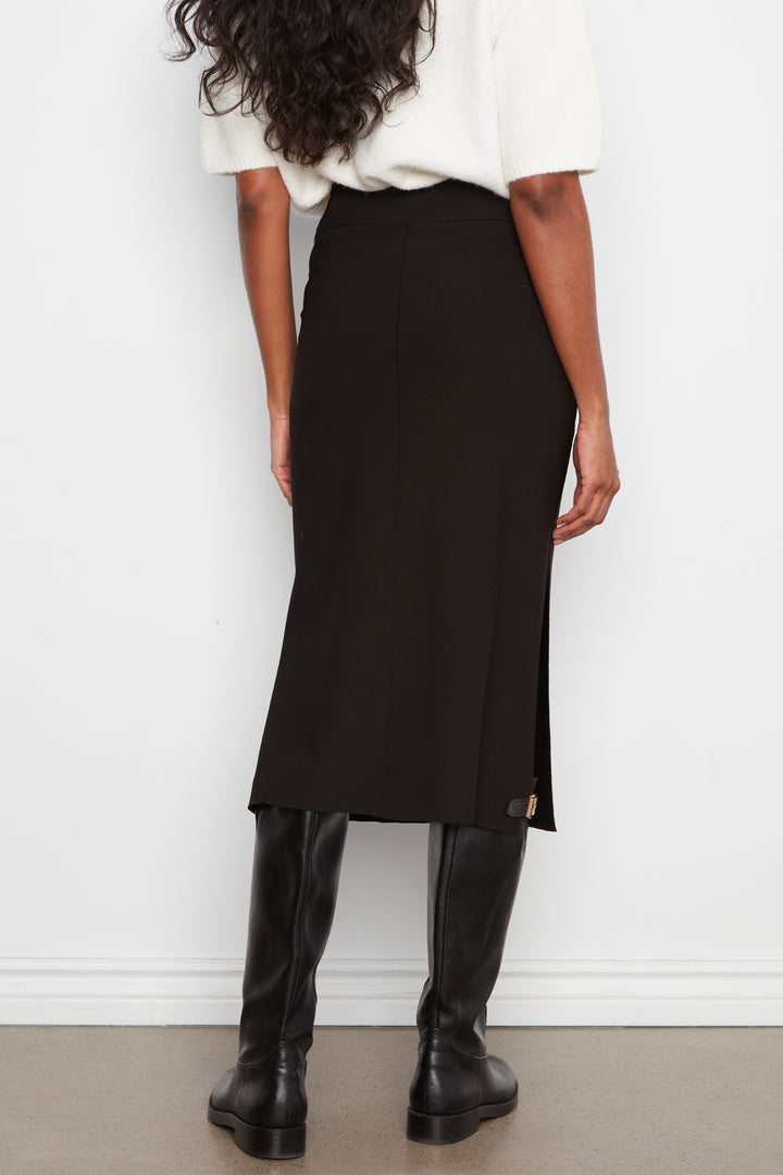 A woman shown from the back wearing a black midi skirt with a side slit, paired with a white top and black knee-high boots, standing against a white wall.