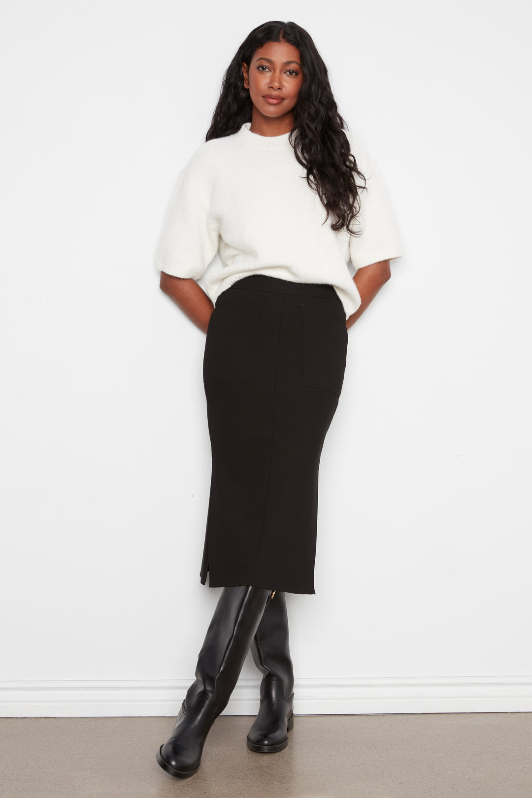 A woman modeling a white short-sleeve knit top and a black midi pencil skirt, paired with black knee-high boots, standing against a plain white wall.