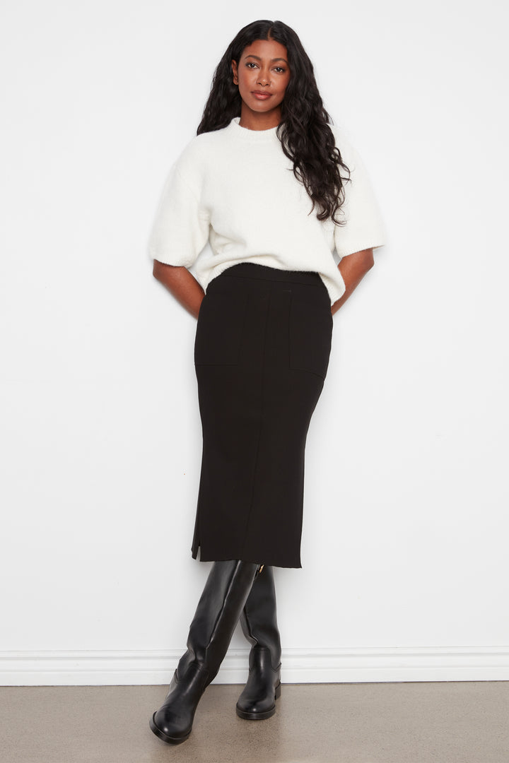 A woman modeling a white short-sleeve knit top and a black midi pencil skirt, paired with black knee-high boots, standing against a plain white wall.