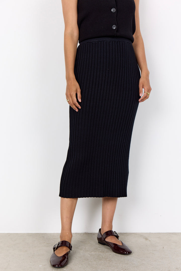 A model wearing a black ribbed knit midi skirt with a matching black sleeveless top and dark brown Mary Jane flats, standing against a white wall.
