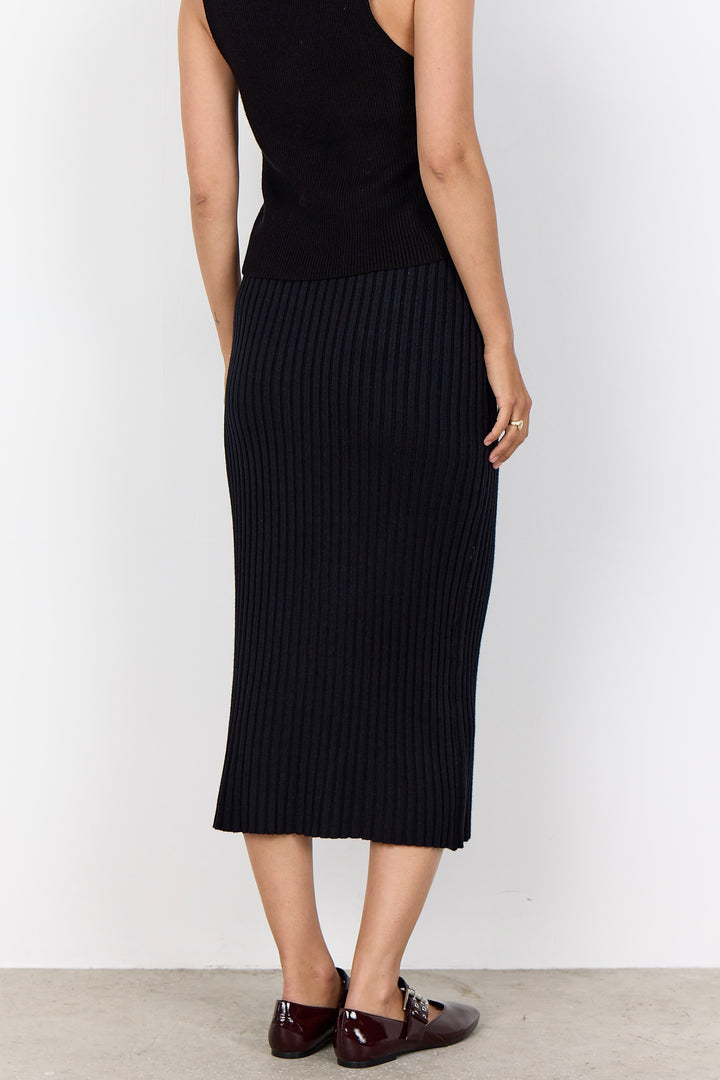 A model from the back wearing a black ribbed knit midi skirt with a black sleeveless top and dark brown leather loafers, set against a plain white wall.
