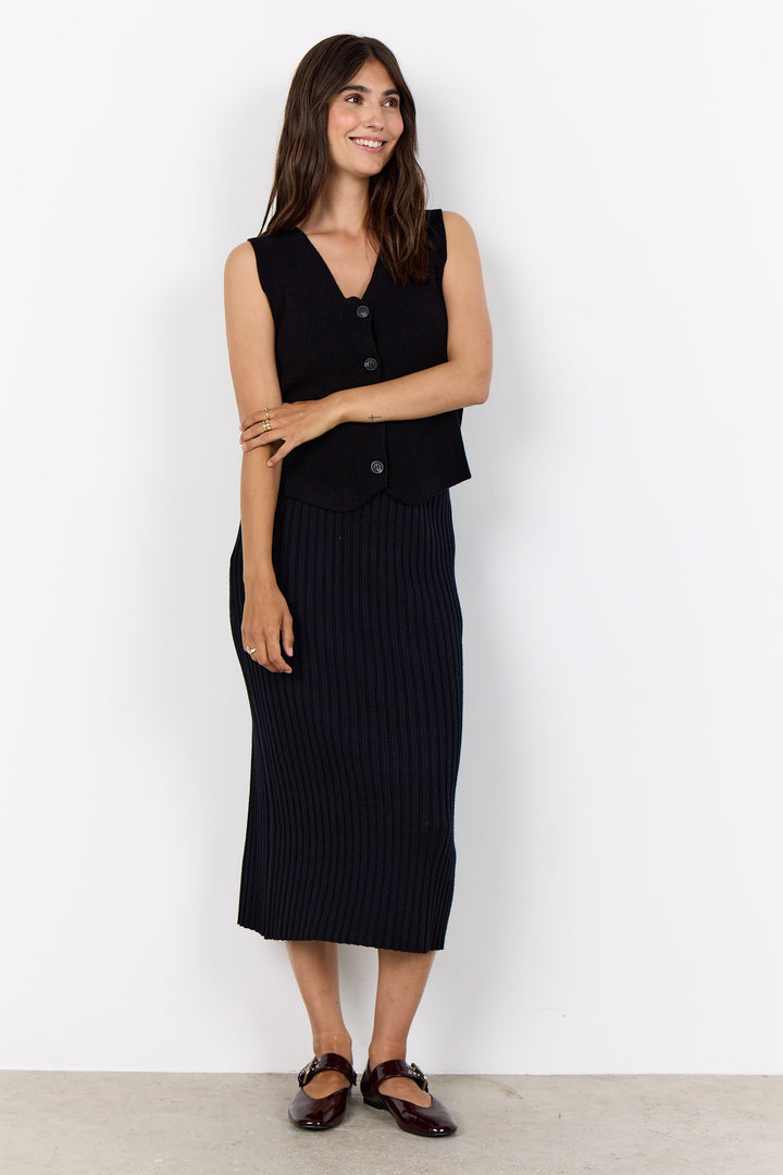 A woman modeling a black sleeveless vest and matching black ribbed midi skirt, paired with dark brown flat shoes, against a plain white wall.