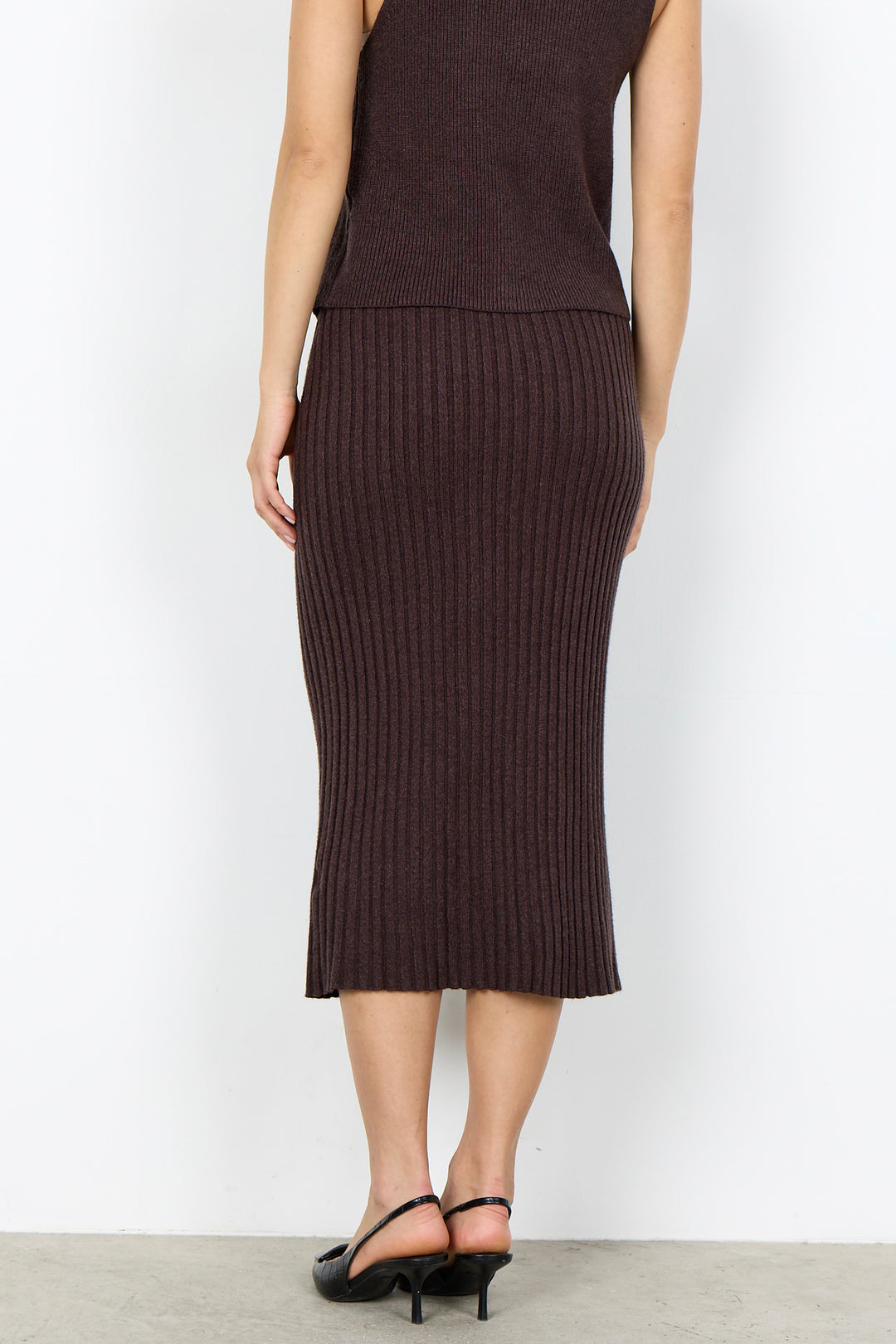 A model from the back wearing a dark brown ribbed knit midi skirt and a matching sleeveless top, paired with black high-heeled sandals, against a plain white studio background.