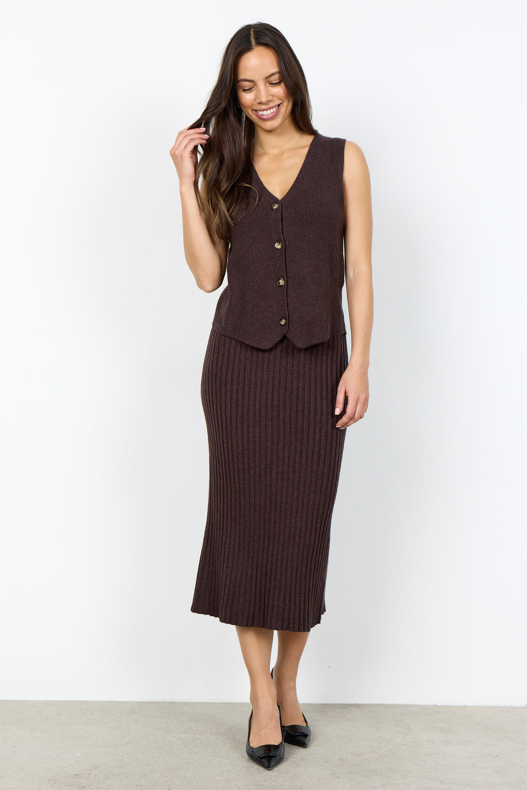 A woman modeling a dark brown ribbed knit vest and midi skirt set against a plain white studio background.